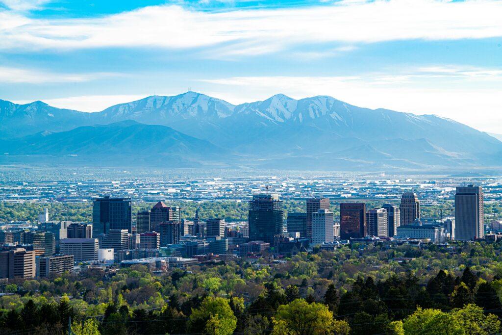 A zoomed-out view of Salt Lake City's skyline, with dense foliage in the foreground and mountains in the background.