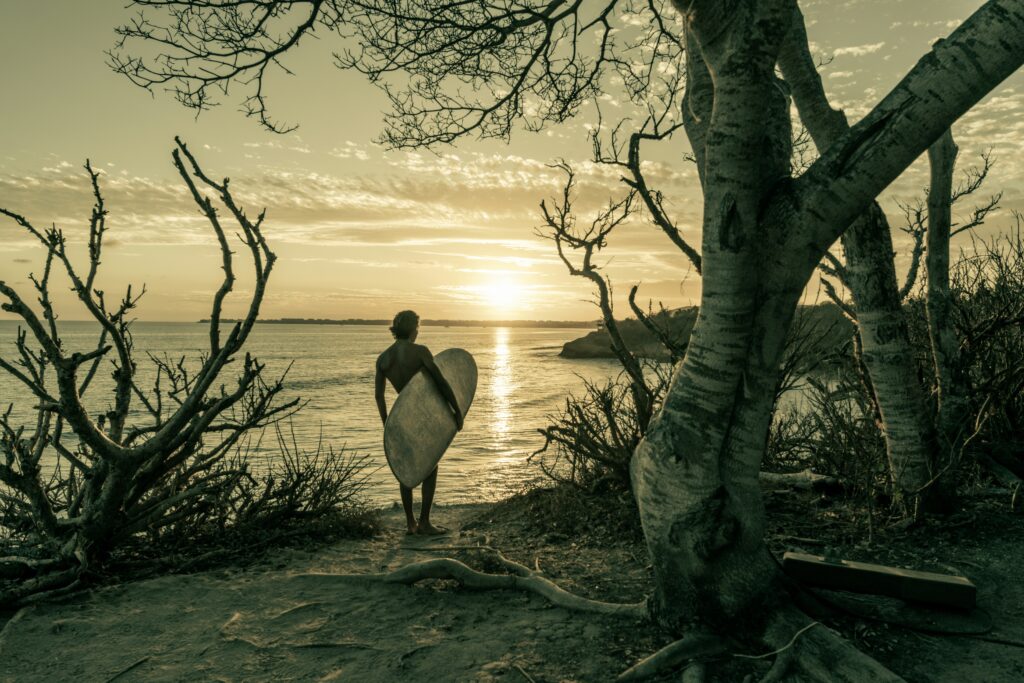 A man with a surfboard walking towards the oceans on a beach filled with driftwood in Sayulita, Mexico.